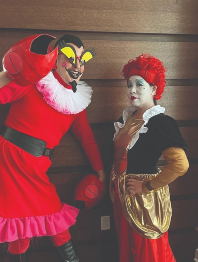 Two people in theatrical costumes and makeup posing indoors against a wood wall, one in bright red and one with a red wig and gold skirt.