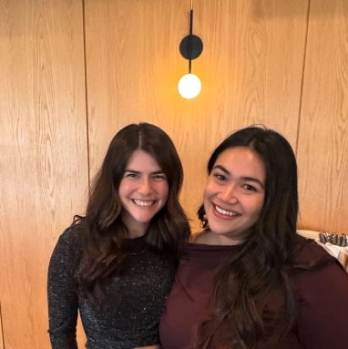Two people smiling together indoors in front of a wood wall with a single round light above them.