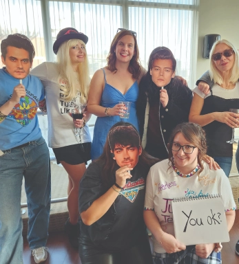 Group photo indoors in front of large windows; everyone is dressed up as different Taylor Swift eras; one person holds a sign that reads “You ok?”.