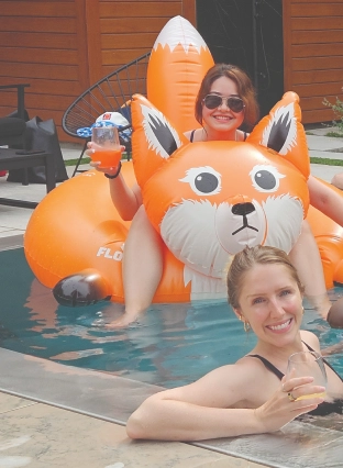 Two people in a swimming pool with a large orange inflatable animal float; one person holds a drink and smiles at the camera.