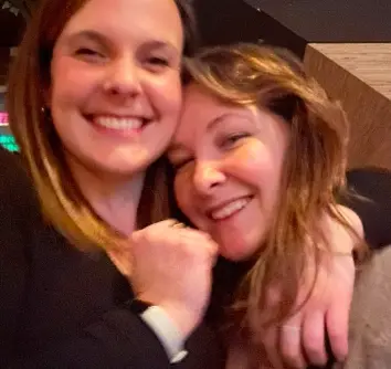 Two coworkers hugging and smiling for a close-up photo indoors.